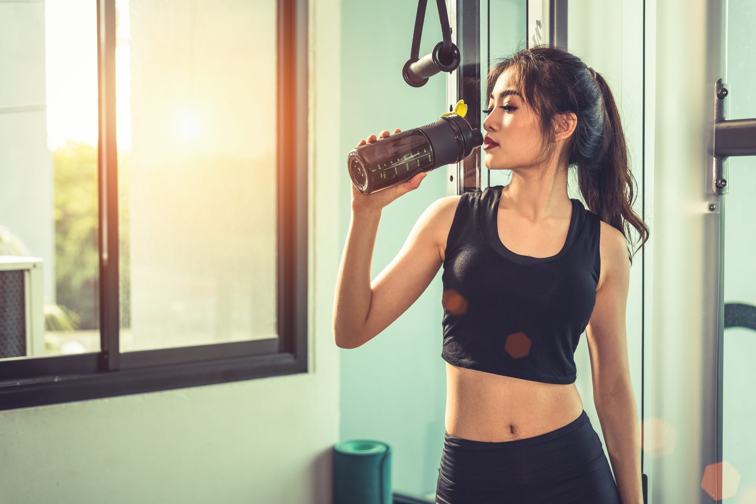 Asian young woman drinking protein shake or water after exercises at fitness gym. Strength training and muscular. Beauty and Healthy concept. Relax concept. Sport equipment and Club center theme.